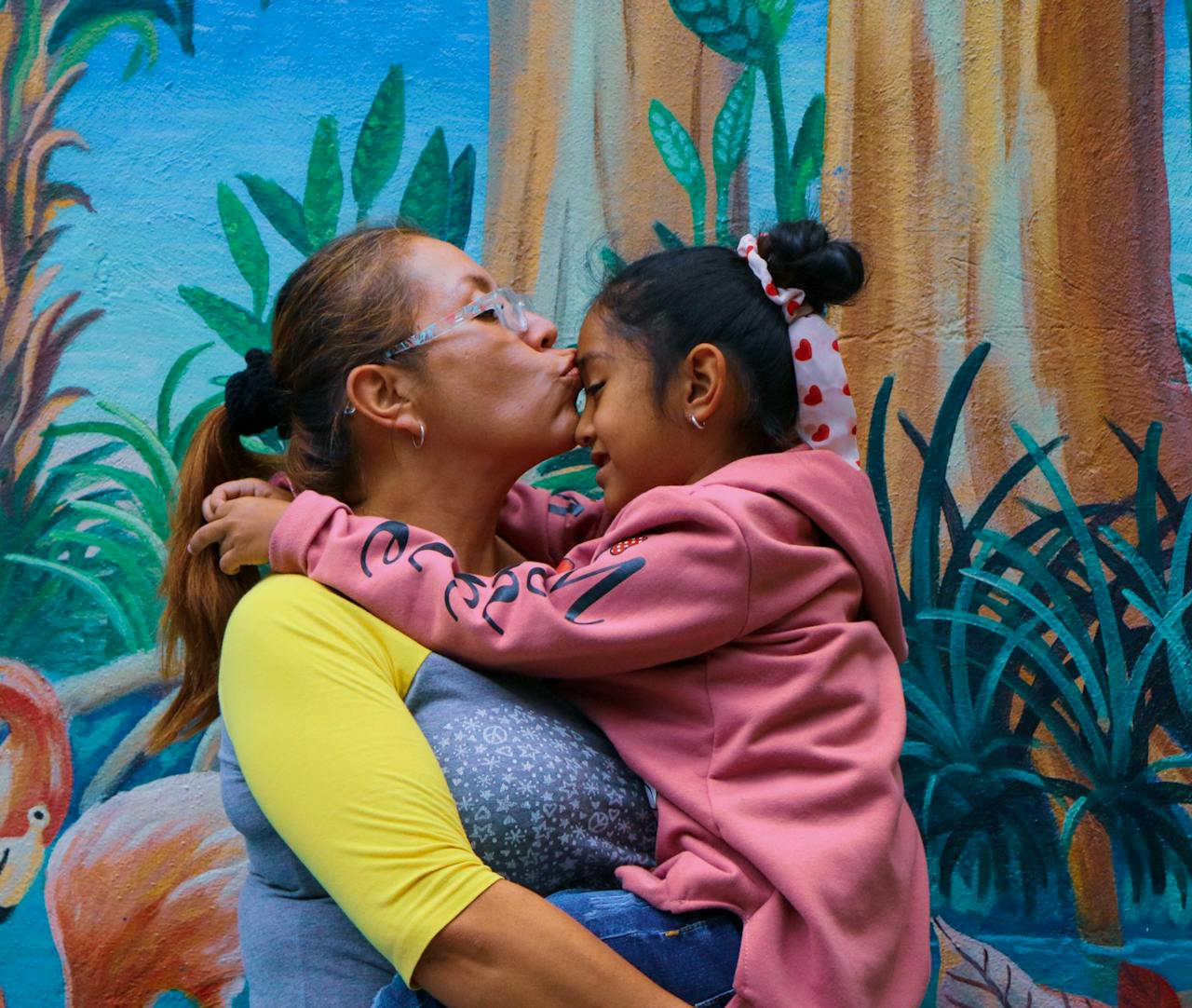 A mother lovingly kisses her daughter outdoors against a colorful mural.