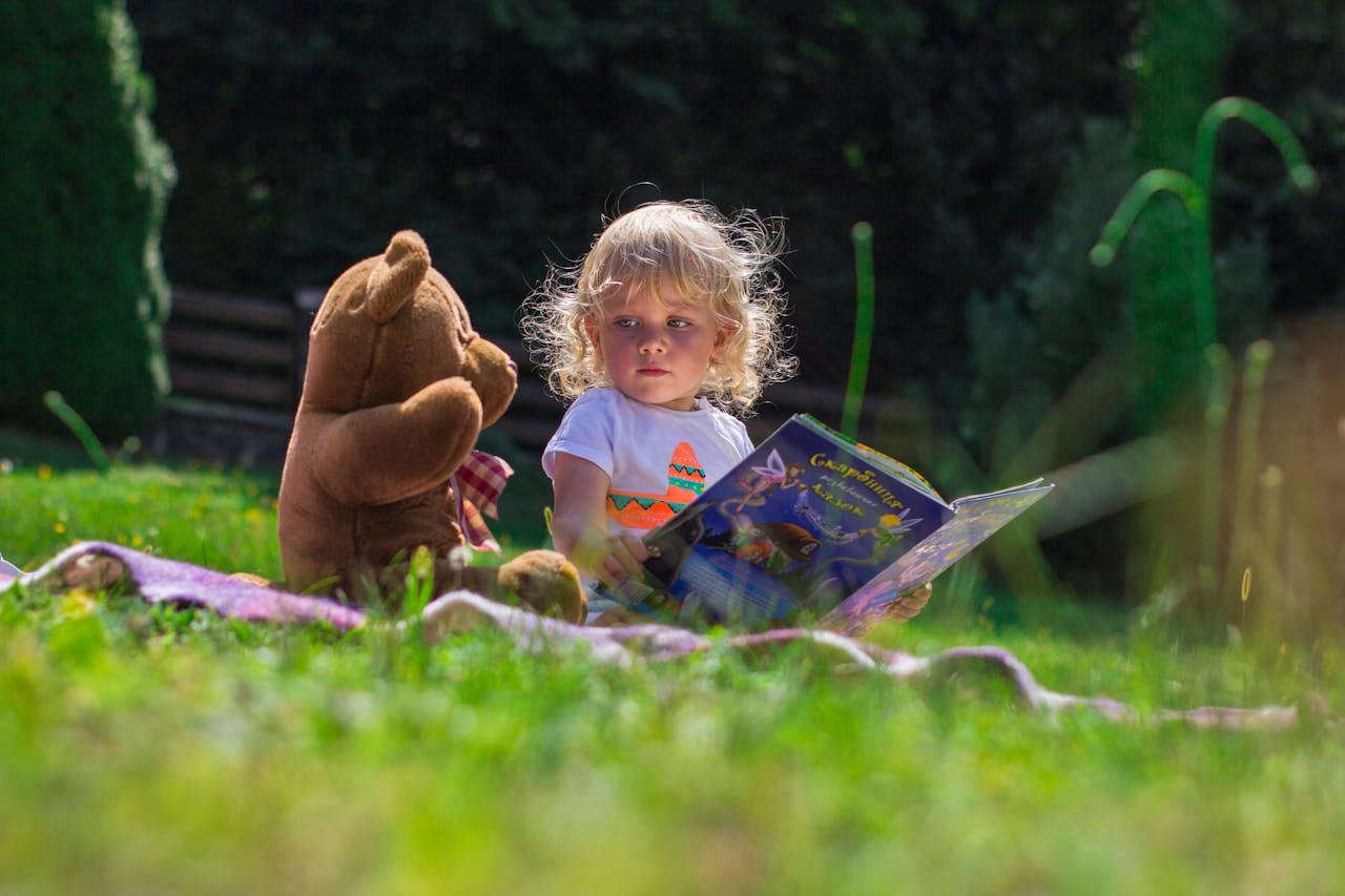 Young child reading a book outdoors with a teddy bear, enjoying a sunny day on the grass.